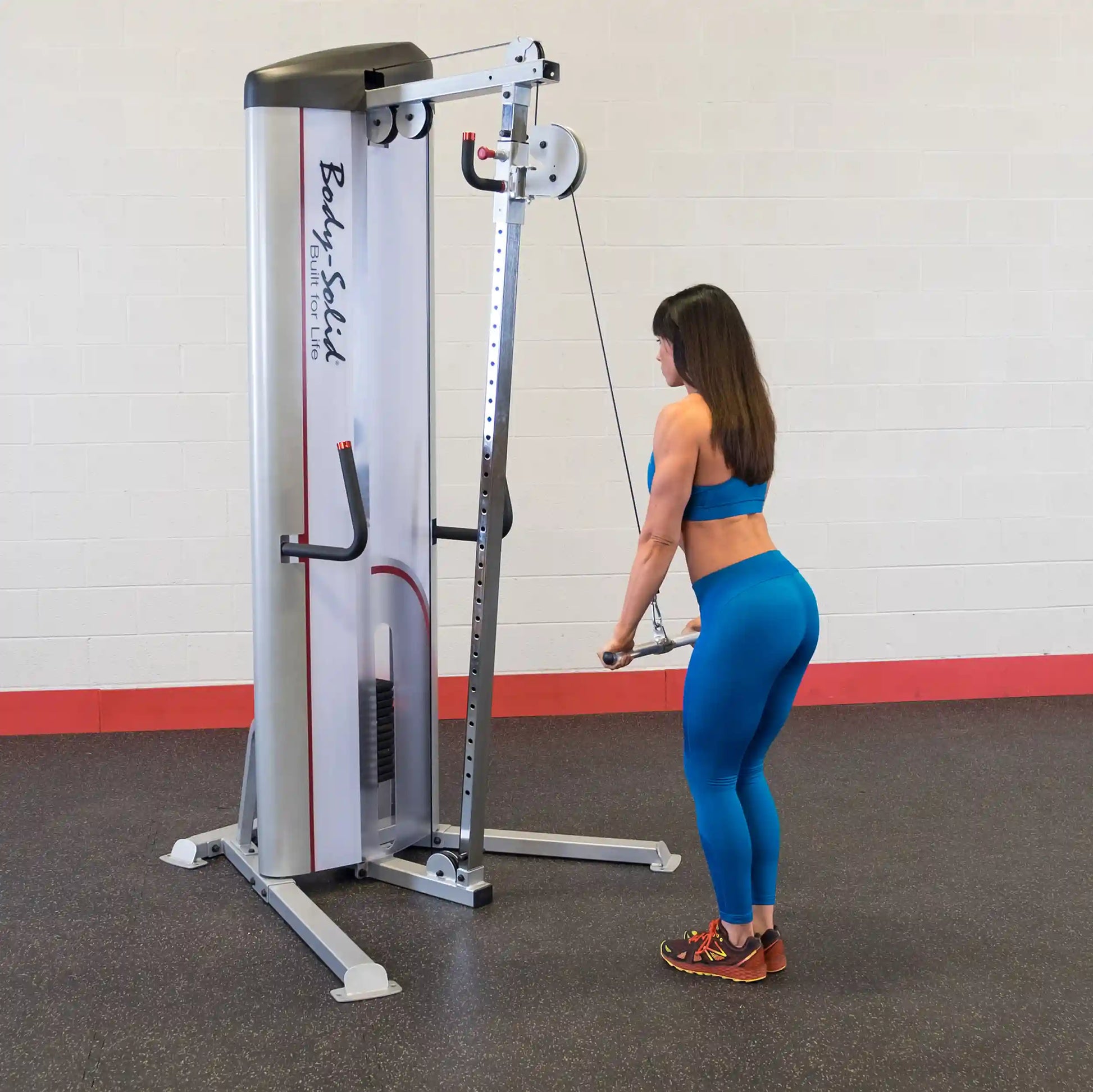 A woman in a blue workout outfit is using the Body-Solid Pro ClubLine Series 2 Cable Column S2CC for exercise, standing with her back to the camera while pulling a cable attachment. The gym has a simple background with a red base.
