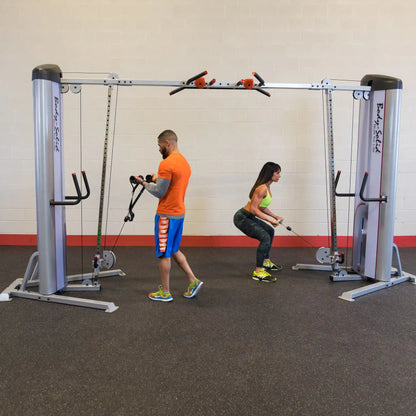 Two individuals exercising with the Body-Solid Pro ClubLine Series 2 Cable Crossover S2CCO, one performing a squat while using the cable machine and the other adjusting the cables for a workout.