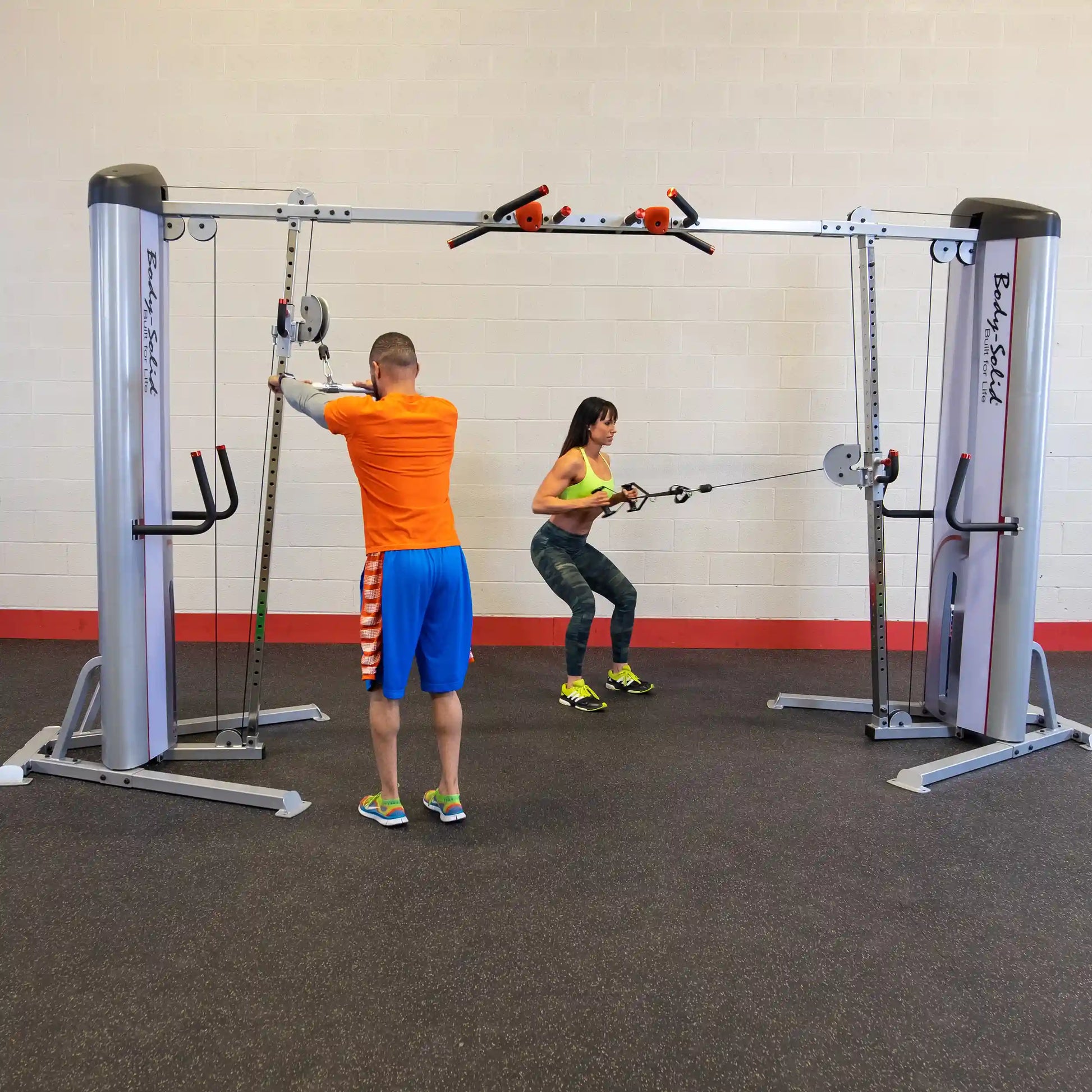 Two individuals exercising with the Body-Solid Pro ClubLine Series 2 Cable Crossover S2CCO in a gym setting, one performing a squat while pulling cables and the other adjusting the equipment.
