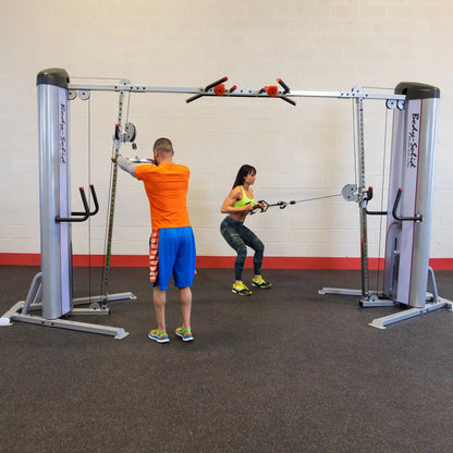 Two individuals exercising with the Body-Solid Pro ClubLine Series 2 Cable Crossover S2CCO in a gym setting, one performing a squat while pulling cables and the other adjusting the equipment.