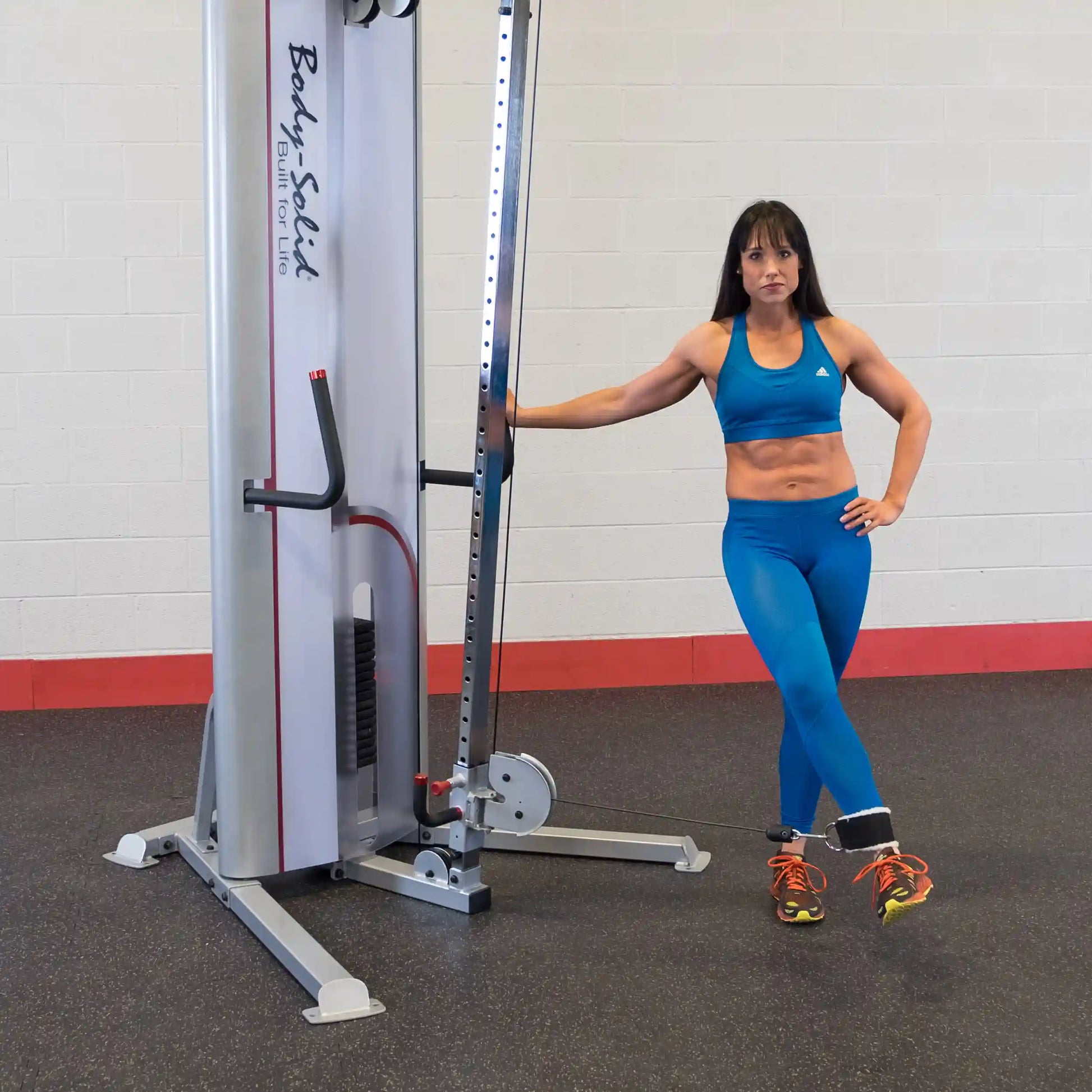 A woman in a blue athletic outfit demonstrating a pose next to the Body-Solid Pro ClubLine Series 2 Cable Crossover S2CCO in a gym setting.