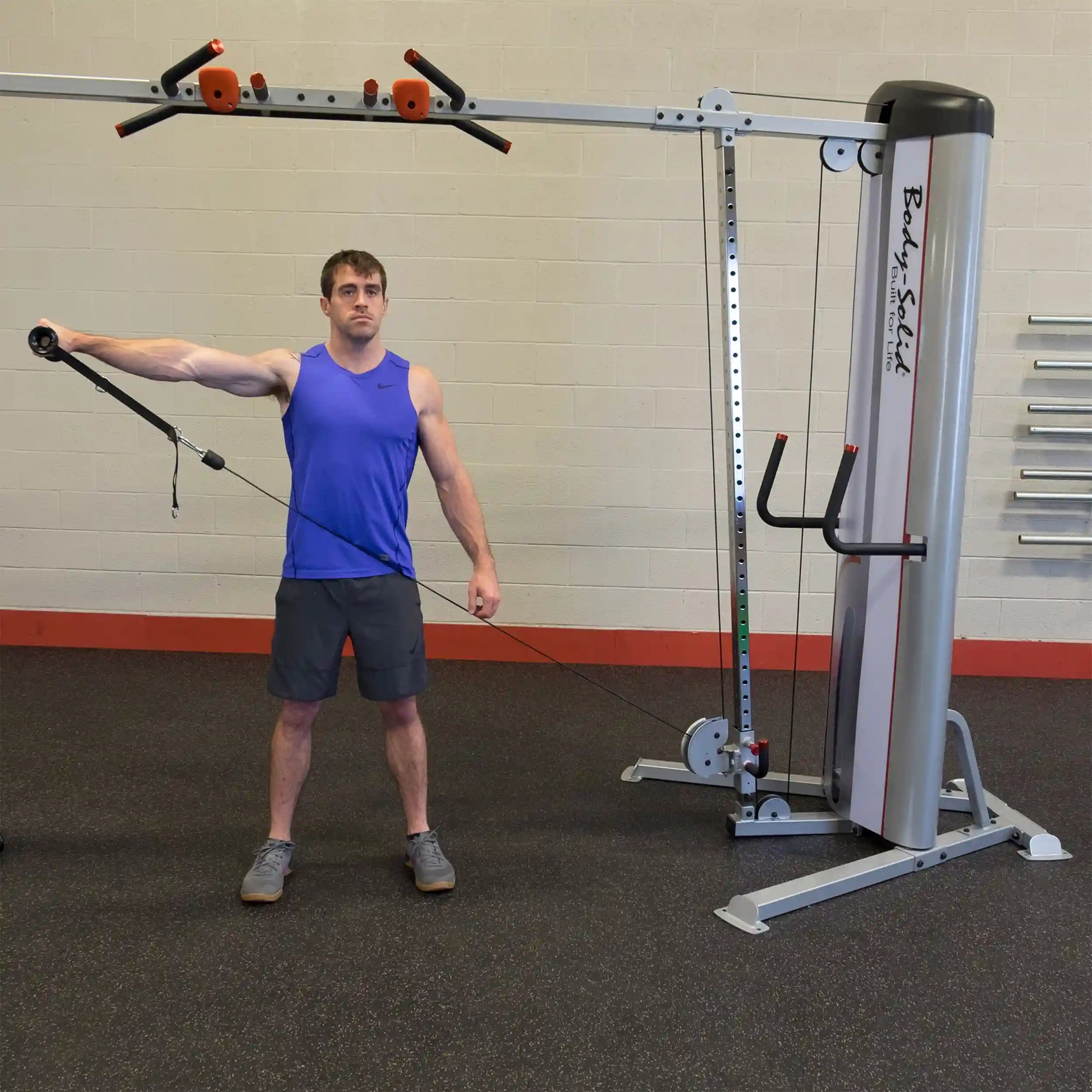 A man in a blue tank top performs an exercise using the Body-Solid Pro ClubLine Series 2 Cable Crossover S2CCO, with one arm extended holding a cable attachment while standing on a gym floor.