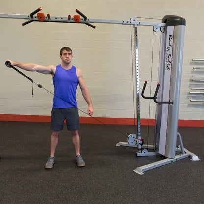 A man in a blue tank top performs an exercise using the Body-Solid Pro ClubLine Series 2 Cable Crossover S2CCO, with one arm extended holding a cable attachment while standing on a gym floor.