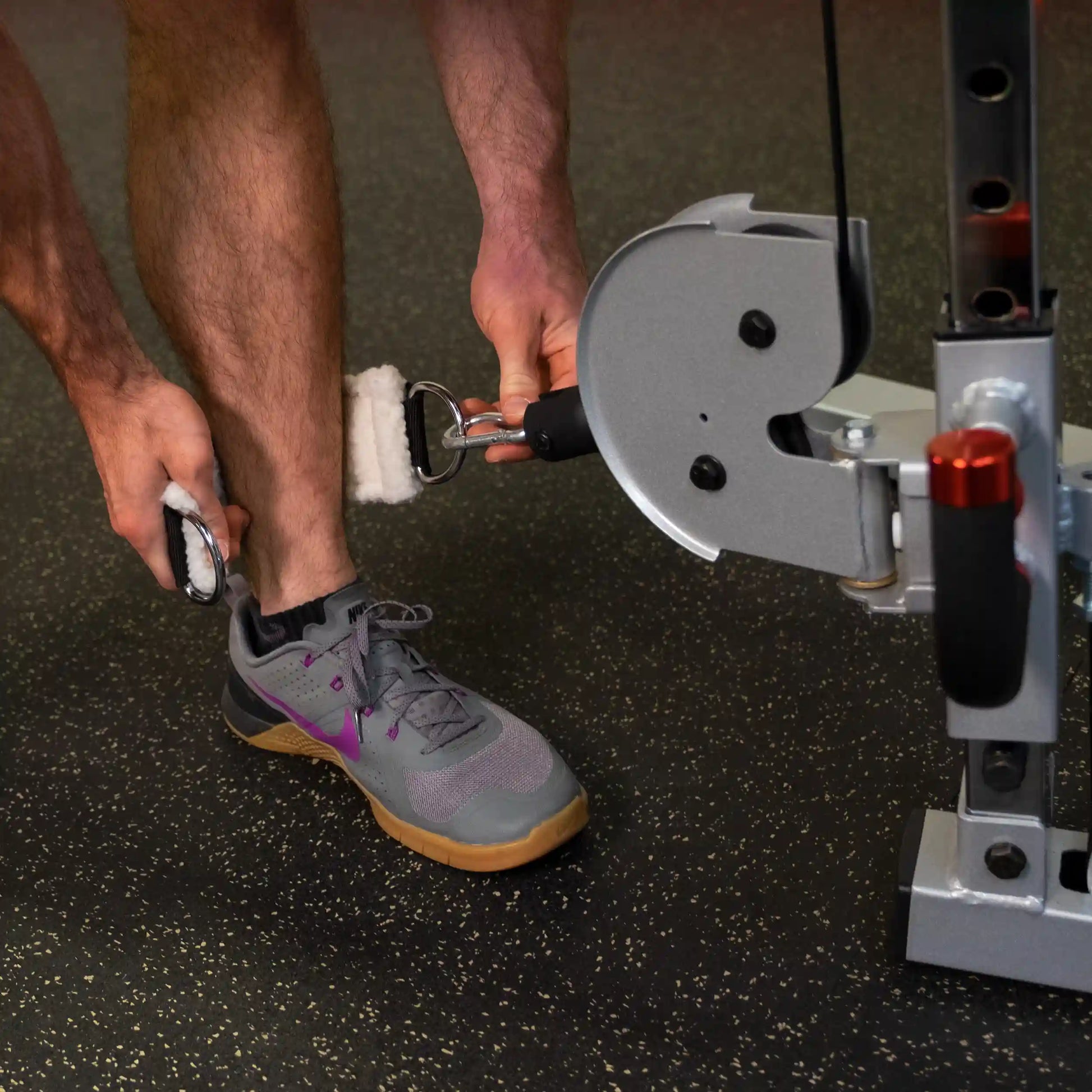 A person using the Body-Solid Pro ClubLine Series 2 Cable Crossover S2CCO exercise equipment, attaching a foot strap to their ankle for a workout.
