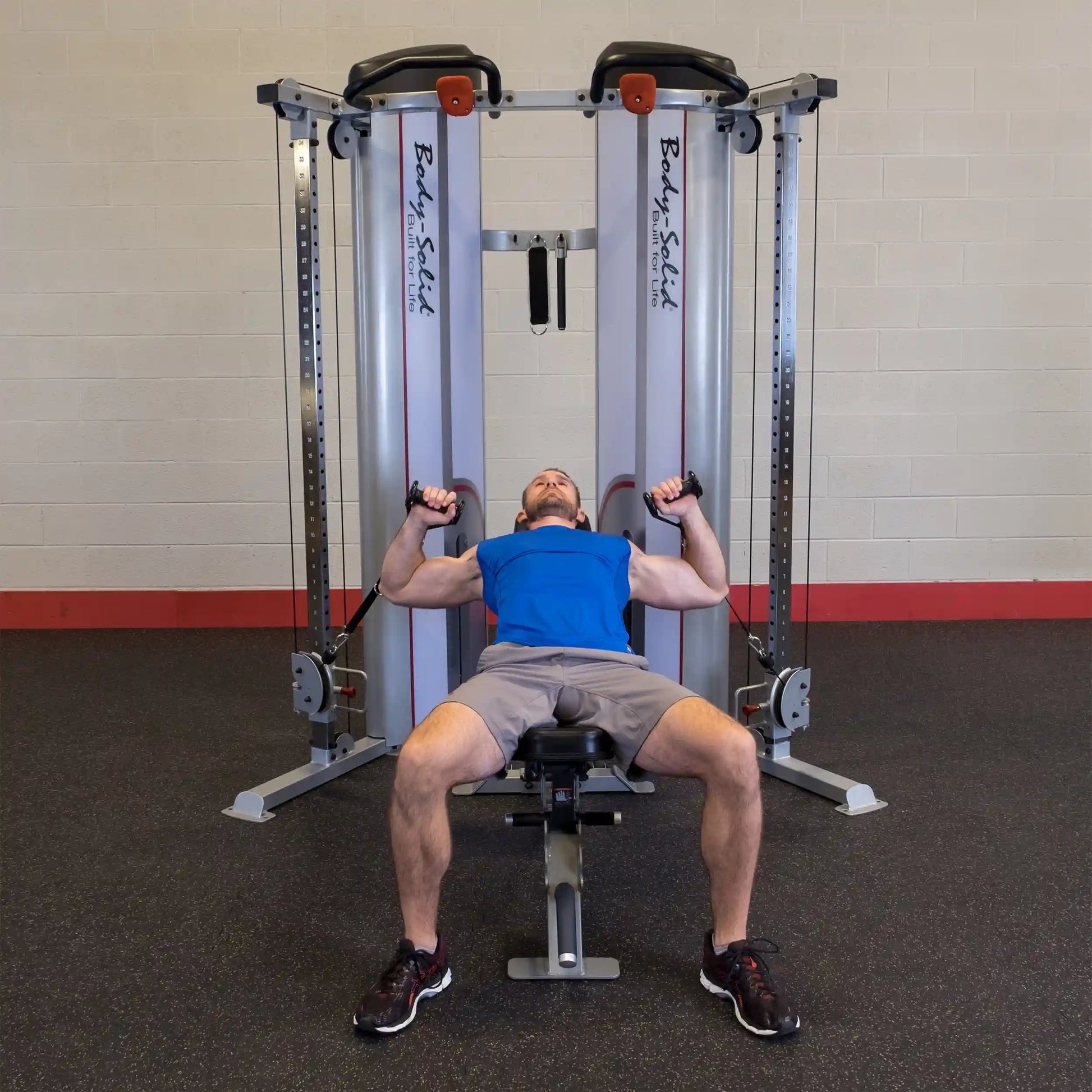 A man performs an exercise on the Body-Solid Pro ClubLine Series 2 Functional Trainer S2FT, lying on a bench with cables in both hands, in a gym setting.