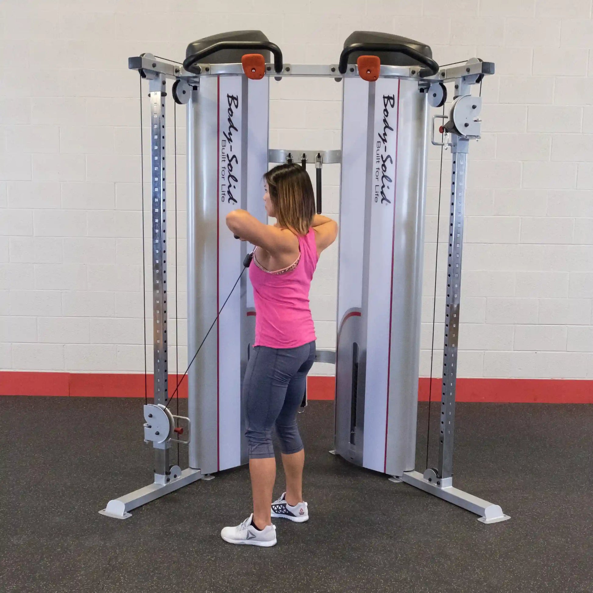 A woman in a pink tank top and gray leggings performs an exercise using the Body-Solid Pro ClubLine Series 2 Functional Trainer S2FT in a gym setting.