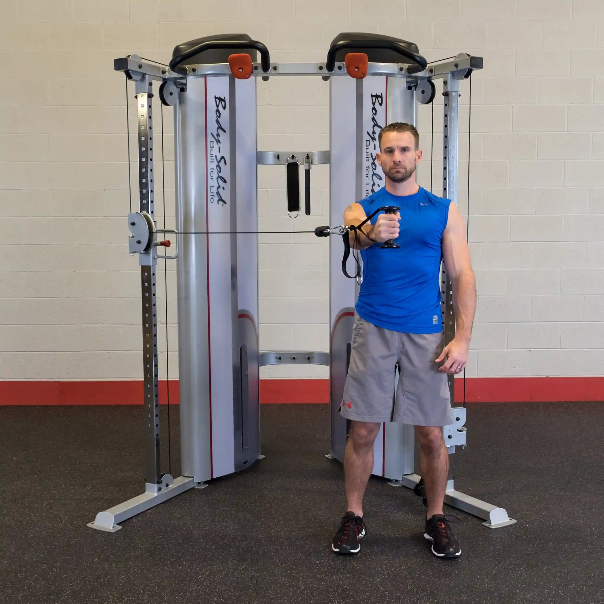 A man in a blue sleeveless shirt demonstrates an exercise using the Body-Solid Pro ClubLine Series 2 Functional Trainer S2FT, standing in front of the equipment with a cable handle in his right hand.