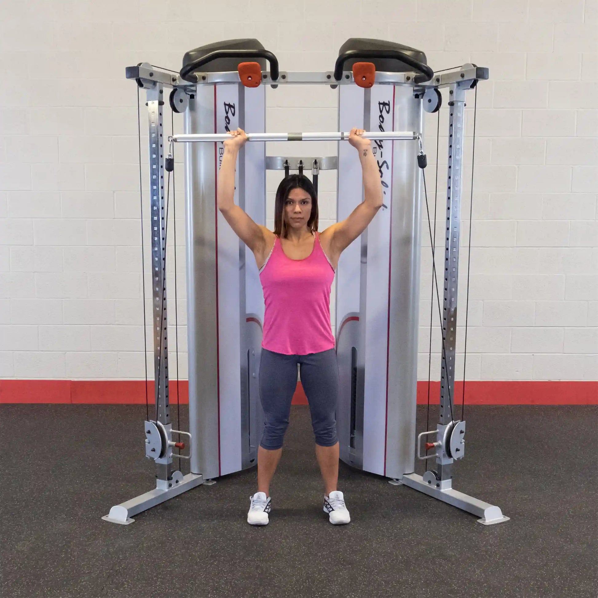 A woman in a pink tank top and gray leggings performs an overhead lift with a barbell in front of the Body-Solid Pro ClubLine Series 2 Functional Trainer S2FT in a gym setting.