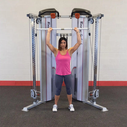 A woman in a pink tank top and gray leggings performs an overhead lift with a barbell in front of the Body-Solid Pro ClubLine Series 2 Functional Trainer S2FT in a gym setting.