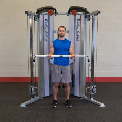 A person stands in front of the Body-Solid Pro ClubLine Series 2 Functional Trainer S2FT, holding a barbell with both hands, wearing a blue sleeveless shirt and gray shorts in a gym setting.