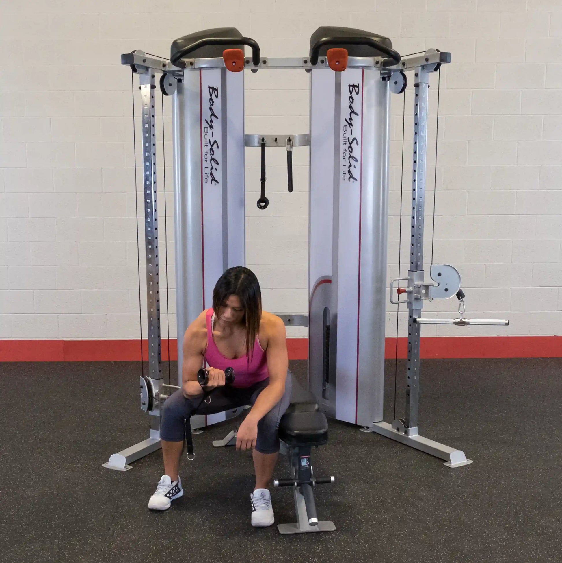 A woman using the Body-Solid Pro ClubLine Series 2 Functional Trainer S2FT, performing a bicep curl while seated on a bench. The gym environment features a gray floor and a white wall with red accents.