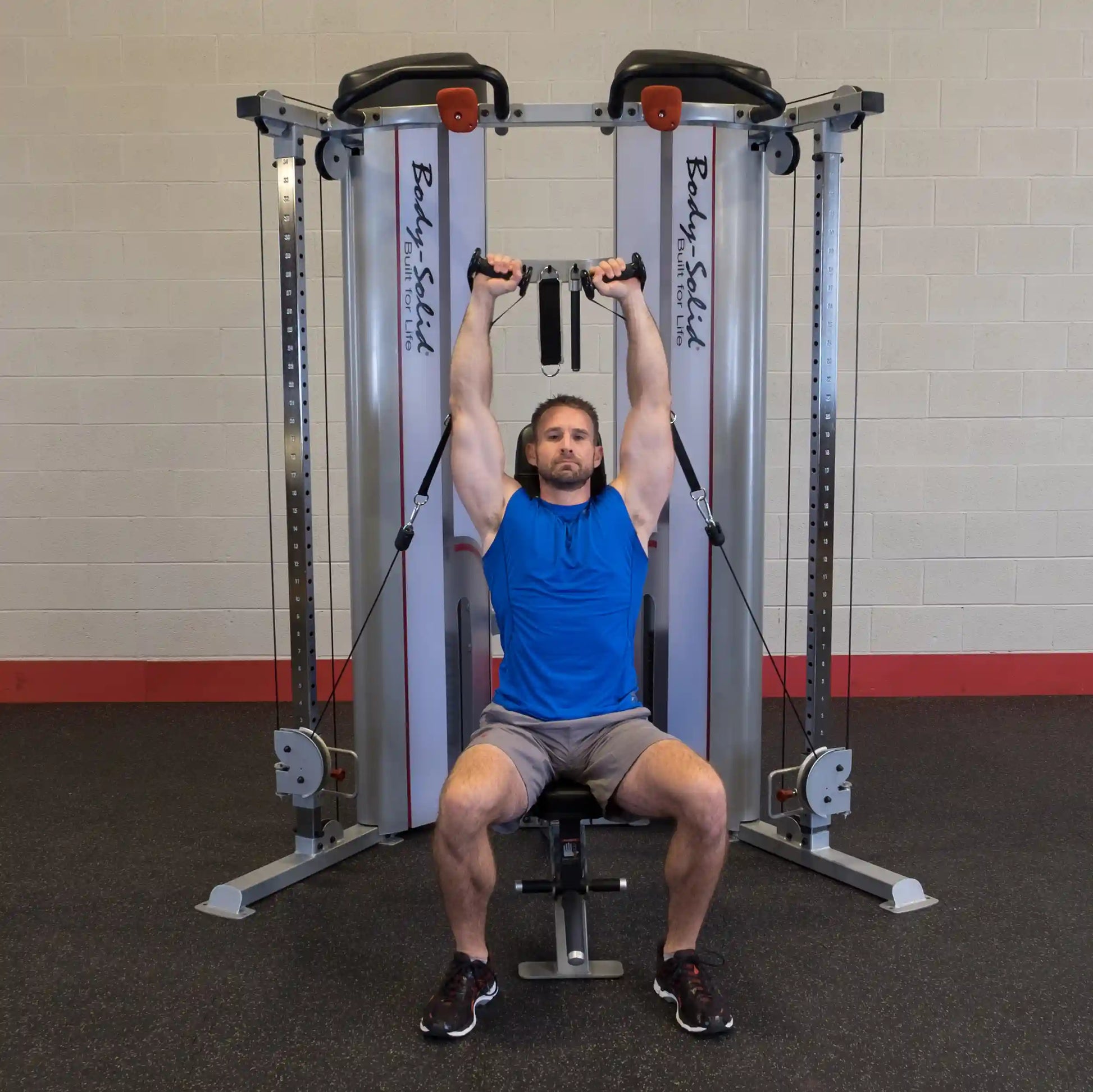 A man performing an overhead exercise using the Body-Solid Pro ClubLine Series 2 Functional Trainer S2FT in a gym setting, seated on a bench with resistance cables attached.