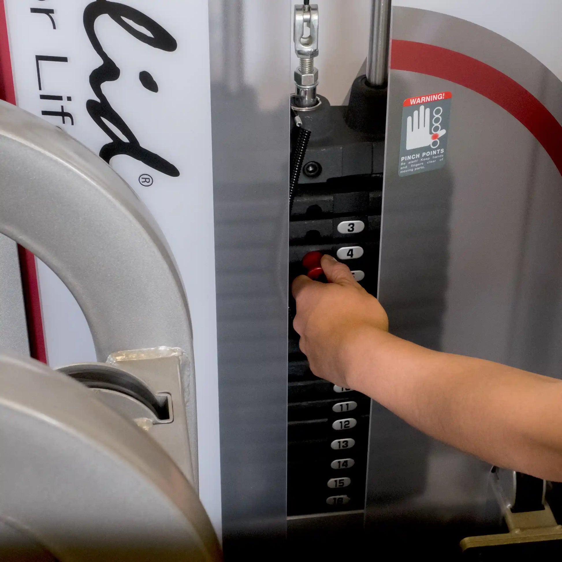 A close-up of a hand adjusting the weight selector on the Body-Solid Pro ClubLine Series 2 Leg Extension & Leg Curl Machine S2LEC. The machine features a warning label about pinch points and a weight stack with numbered plates.