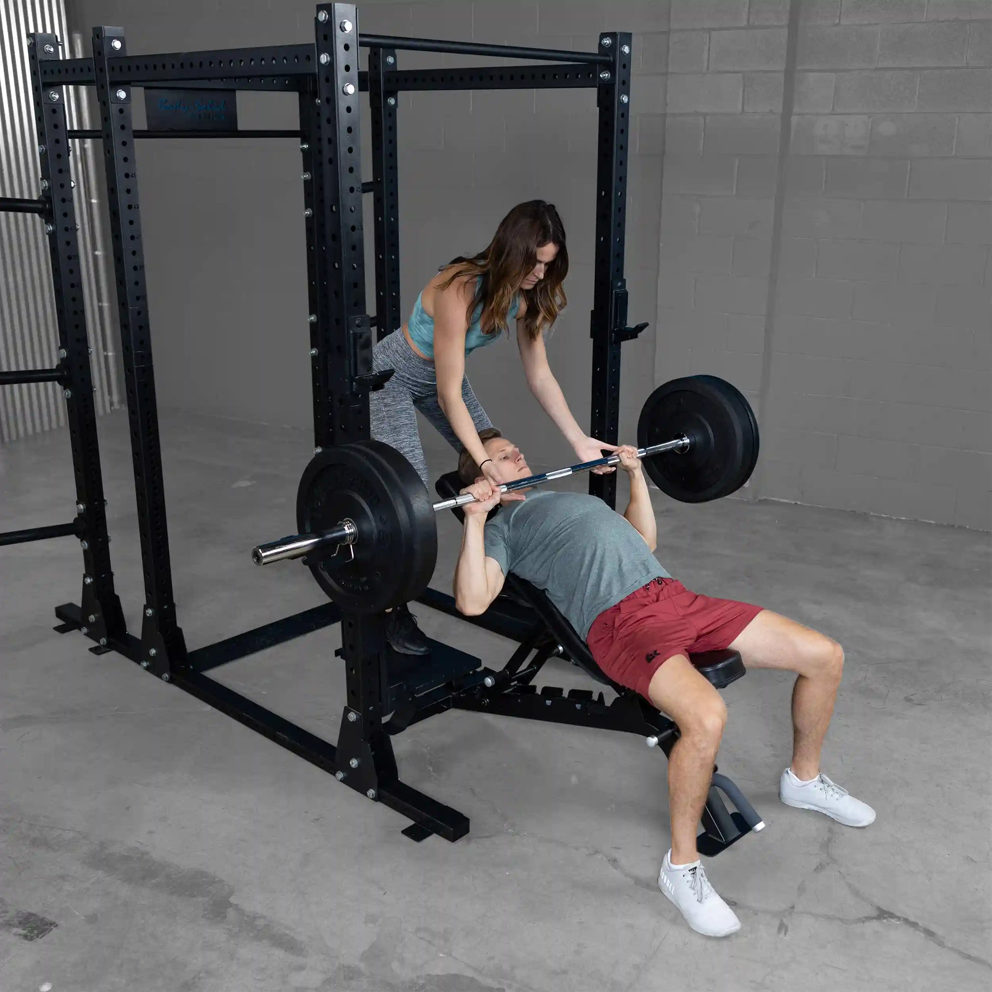 A woman assists a man with a barbell bench press using Body-Solid Pro ClubLine Spotter Platforms SPRSP, in a gym setting with gray walls and a black rack.