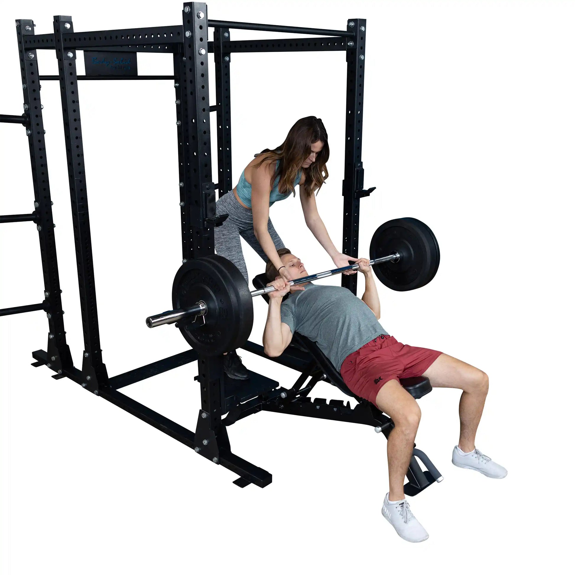 A man performs a bench press with the Body-Solid Pro ClubLine Spotter Platforms SPRSP, assisted by a woman, in a gym setting.
