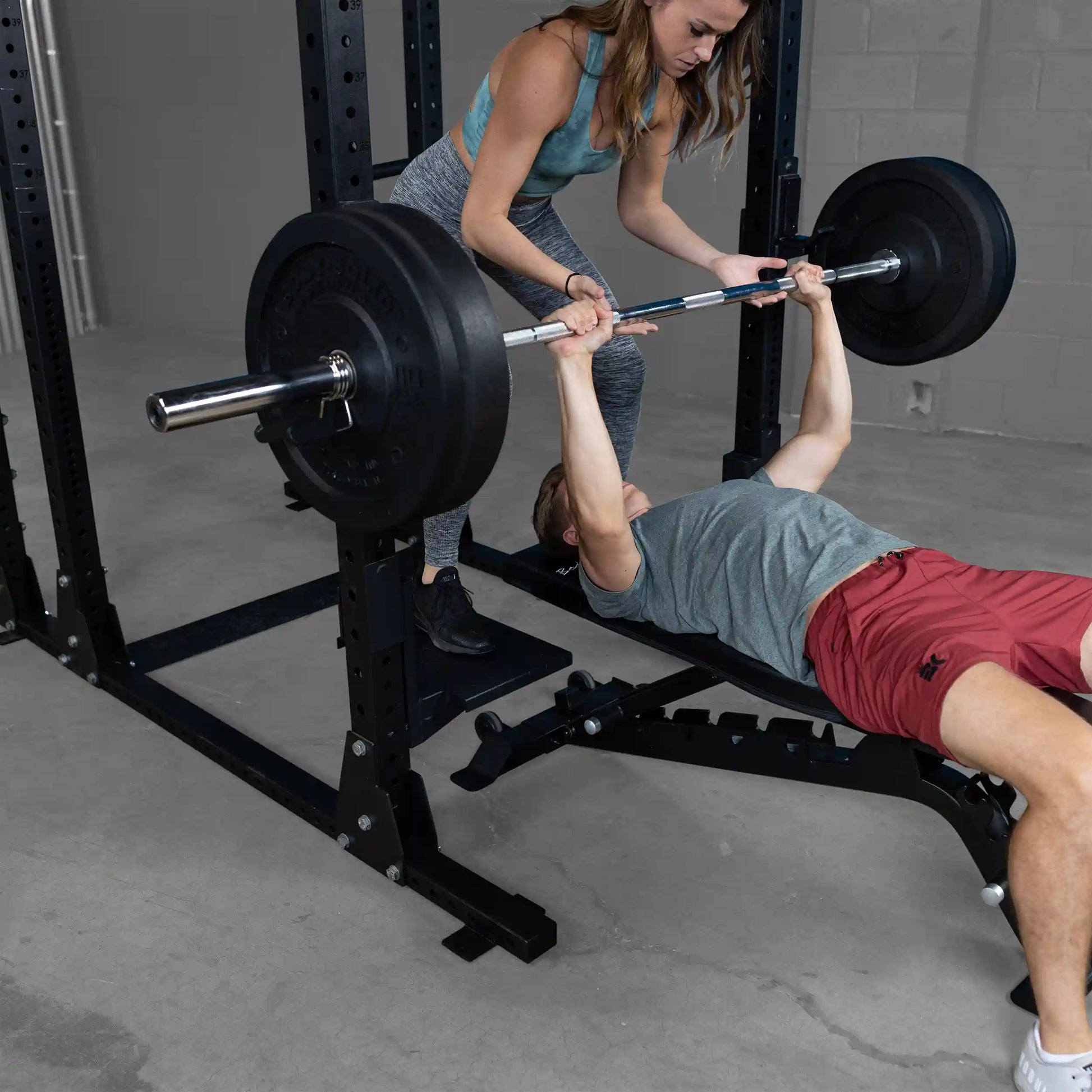 A man performs a bench press with the help of a woman spotting him, using the Body-Solid Pro ClubLine Spotter Platforms SPRSP in a gym setting.