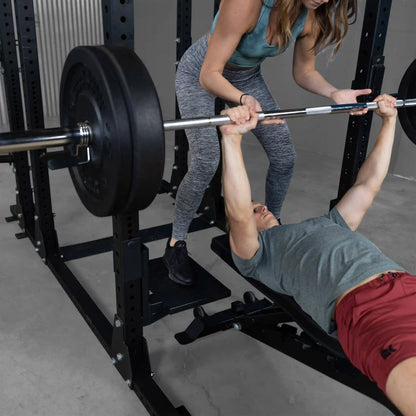 A man performing a bench press with a barbell while being assisted by a woman. The setup includes the Body-Solid Pro ClubLine Spotter Platforms SPRSP in a gym environment.
