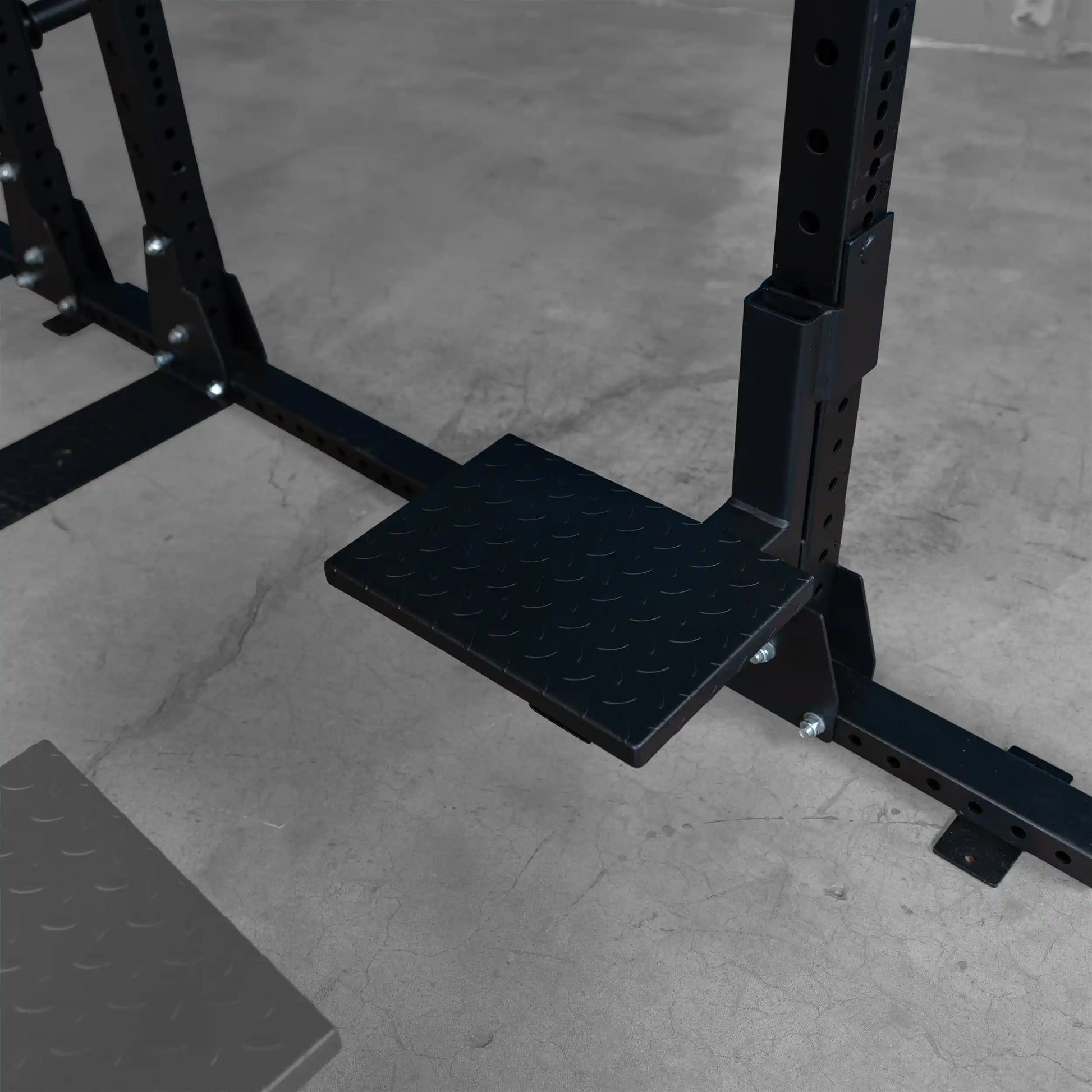 Close-up of the Body-Solid Pro ClubLine Spotter Platforms SPRSP, featuring a sturdy, textured black platform positioned on a gym equipment frame with a concrete floor background.