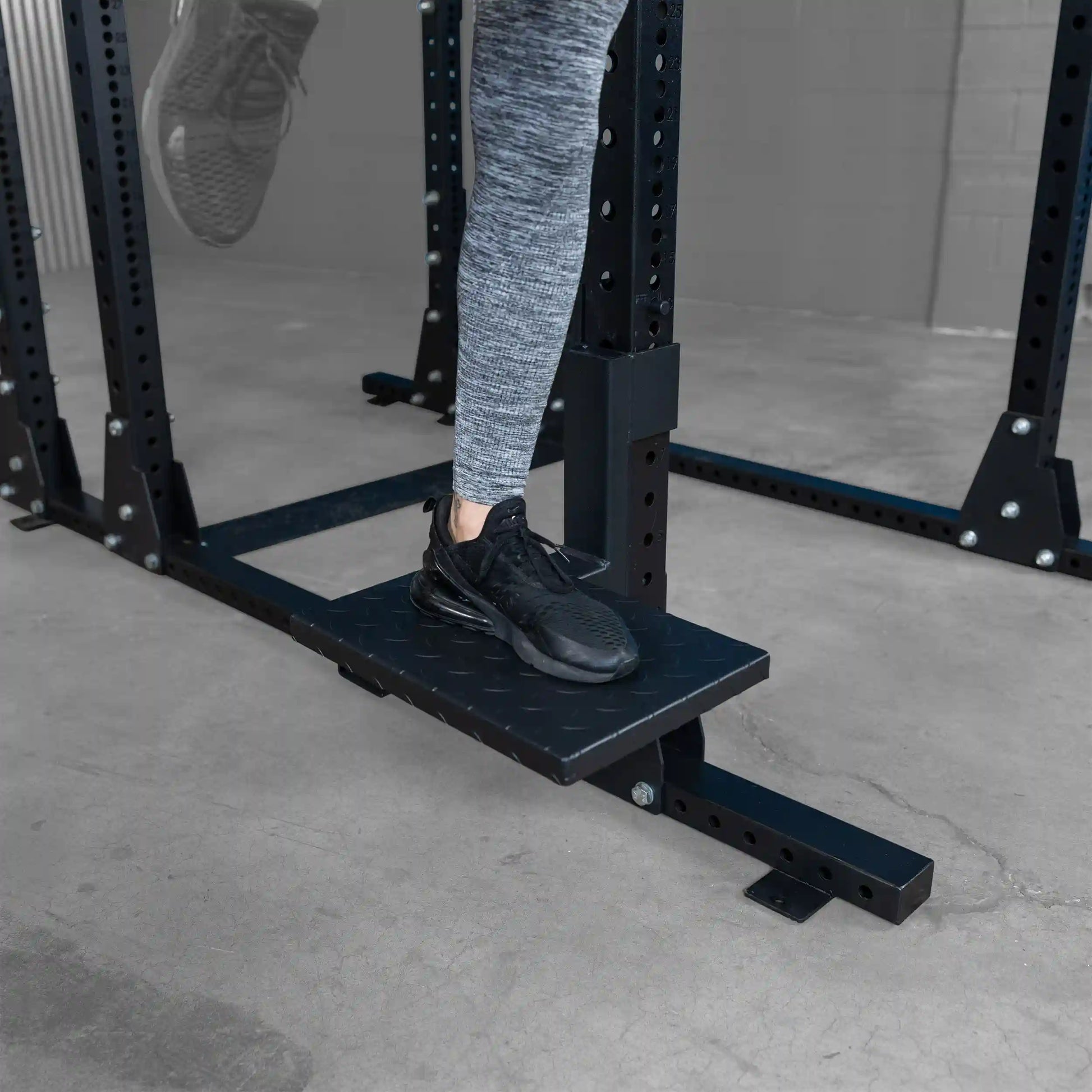 A person wearing gray athletic leggings and black sneakers stands on a Body-Solid Pro ClubLine Spotter Platform SPRSP, which is black and positioned underneath a weightlifting rack in a gym setting.