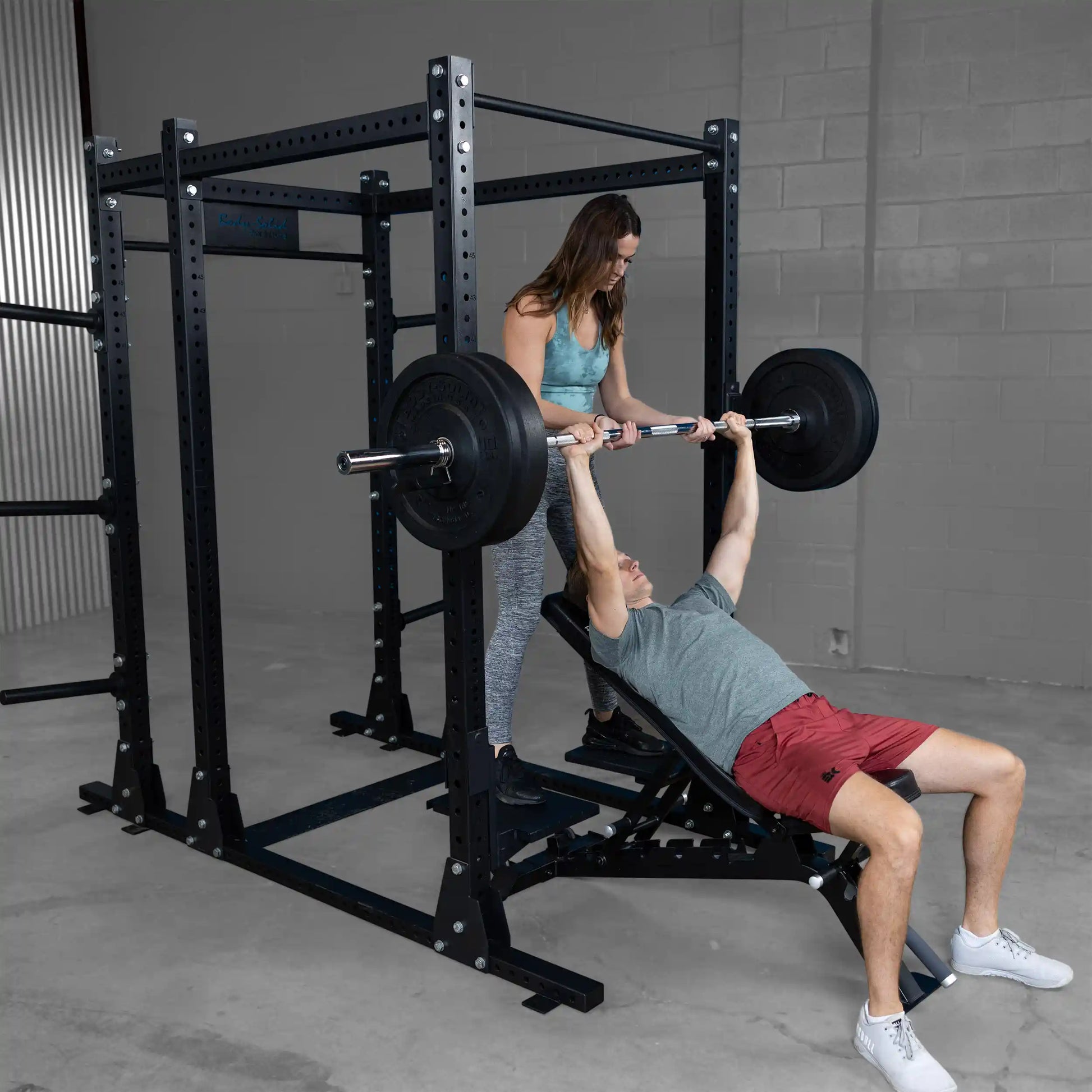 A woman assists a man with a bench press under the Body-Solid Pro ClubLine Spotter Platforms SPRSP, showcasing a well-equipped weightlifting area.