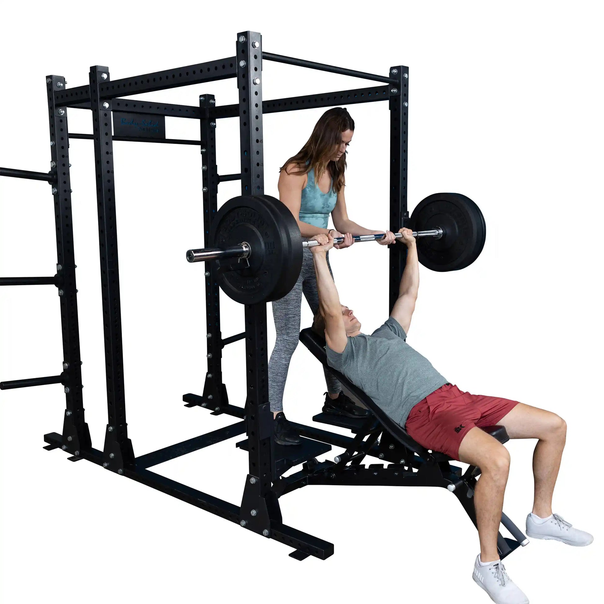 A woman assists a man lifting weights on the Body-Solid Pro ClubLine Spotter Platforms SPRSP, demonstrating a bench press exercise in a gym setting.