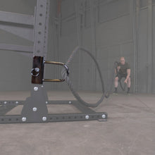 A close-up of the Body-Solid Pro ClubLine U-Link Attachment SPRUL attached to a workout frame, with a person in the background using battle ropes in a gym setting.