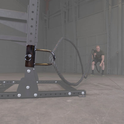 A close-up of the Body-Solid Pro ClubLine U-Link Attachment SPRUL attached to a workout frame, with a person in the background using battle ropes in a gym setting.