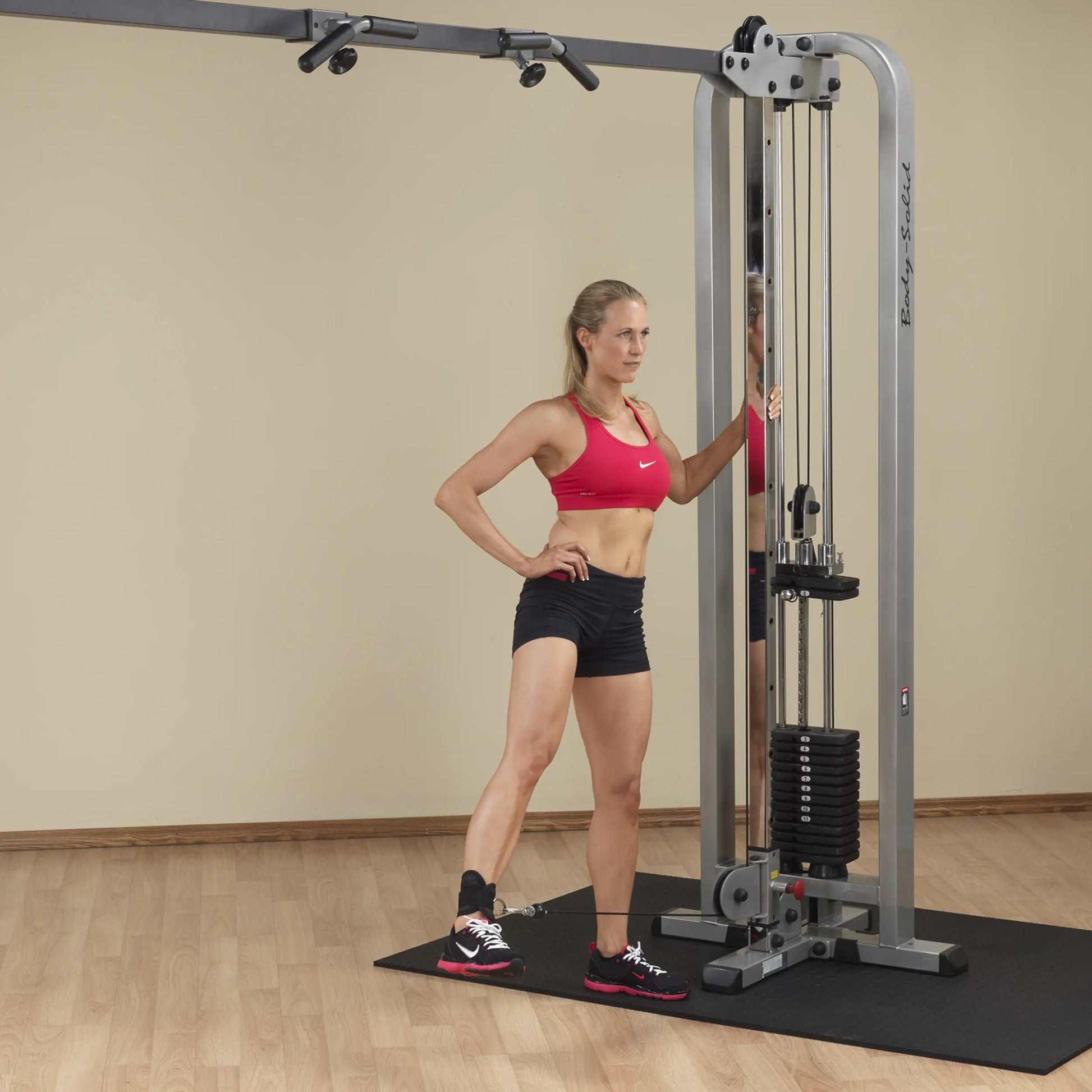 A woman in a red sports bra and black shorts stands beside the Body-Solid Pro Clubline Cable Crossover SCC1200G, demonstrating its use in a fitness setting.