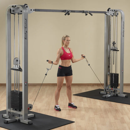A woman performing an exercise with the Body-Solid Pro Clubline Cable Crossover SCC1200G, using cables from both sides while standing on a gym floor.