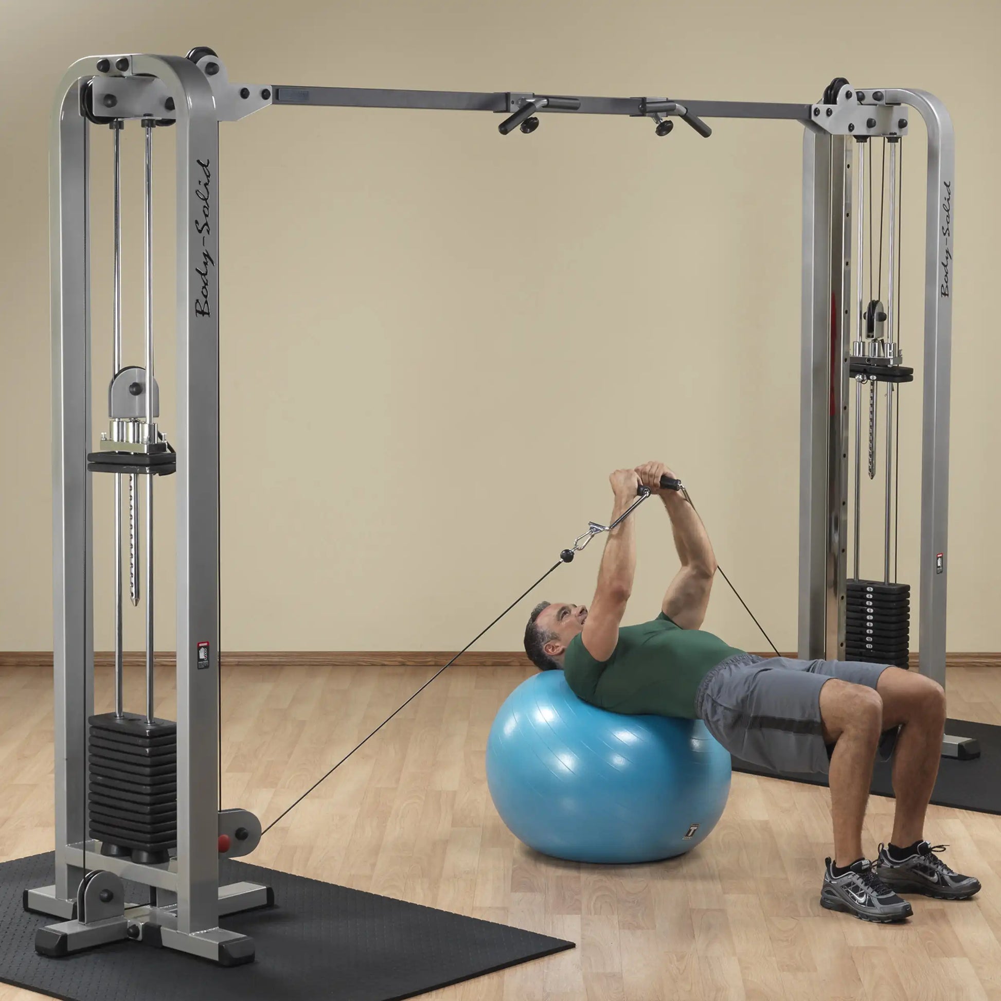 A man performing an exercise on a blue stability ball using the Body-Solid Pro Clubline Cable Crossover SCC1200G, with dual cable pulleys and weight stacks visible in the gym setting.