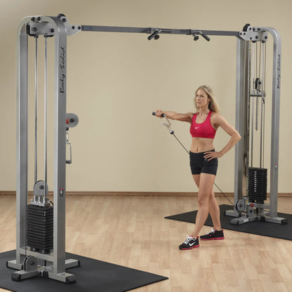 A woman performing an exercise on the Body-Solid Pro Clubline Cable Crossover SCC1200G, showcasing the fitness machine in a gym setting with a wooden floor and neutral background.