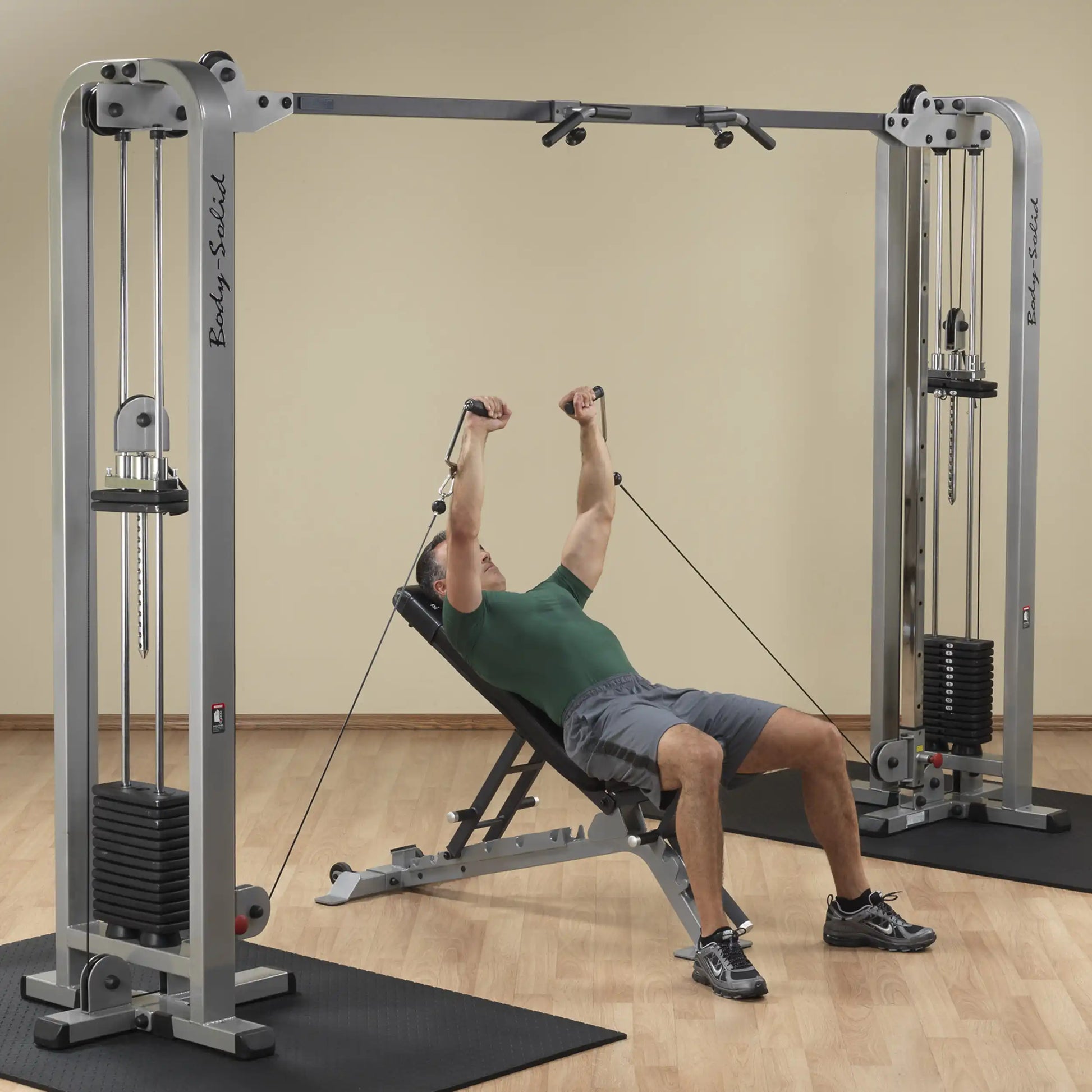 A man performing an exercise on the Body-Solid Pro Clubline Cable Crossover SCC1200G, lying on a bench and using the cable pulleys for resistance training in a gym setting.