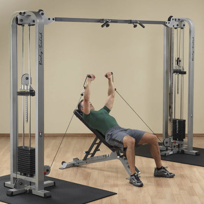 A man performing an exercise on the Body-Solid Pro Clubline Cable Crossover SCC1200G, lying on a bench and using the cable pulleys for resistance training in a gym setting.
