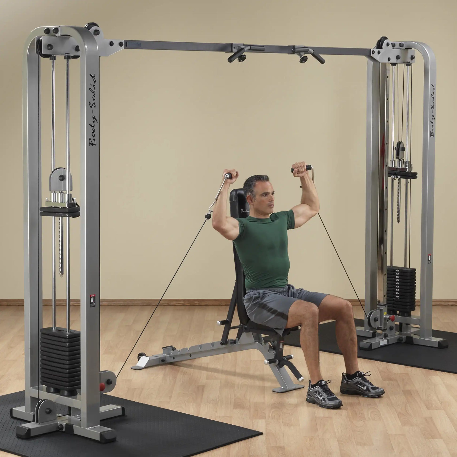 A man using the Body-Solid Pro Clubline Cable Crossover SCC1200G, performing a seated exercise with cables, in a gym setting with wooden flooring.