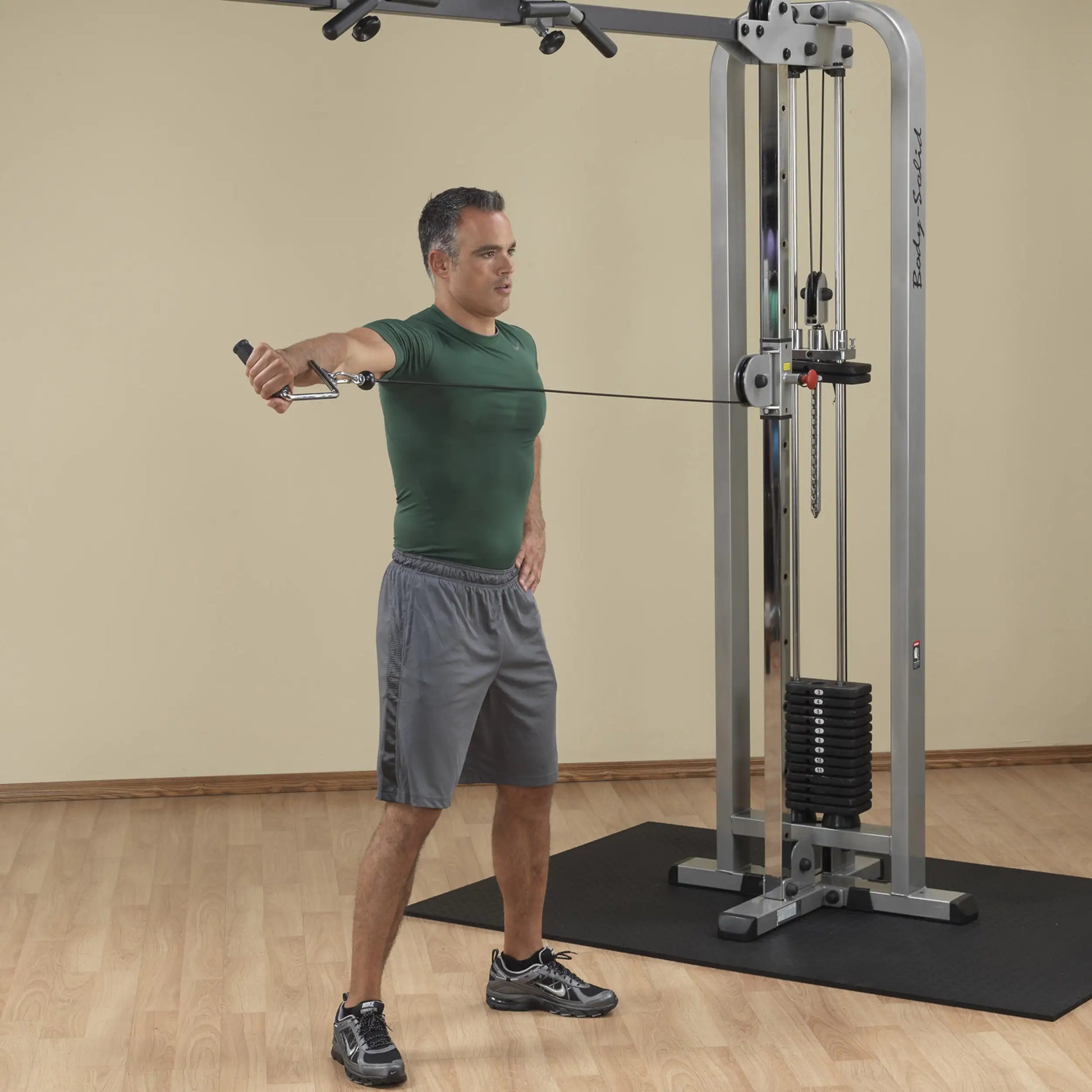 A man in a green shirt and gray shorts performing an exercise using the Body-Solid Pro Clubline Cable Crossover SCC1200G, demonstrating a cable crossover movement in a gym setting.