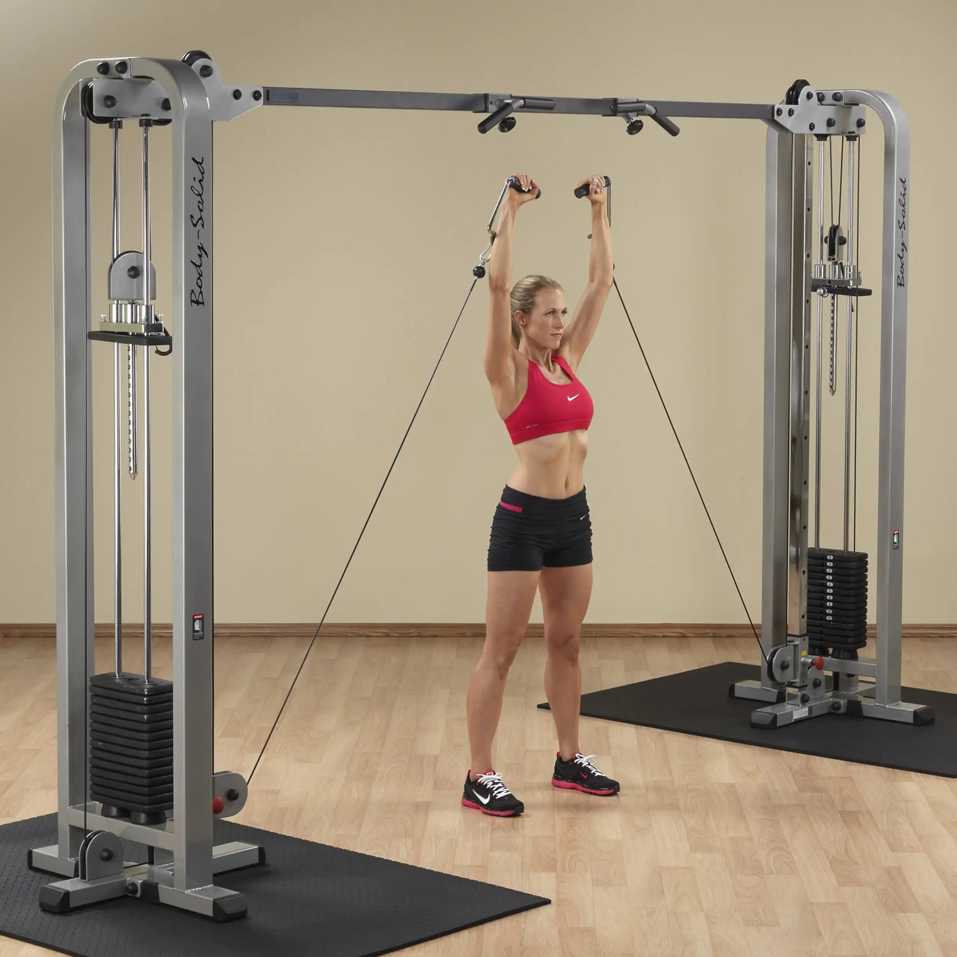A woman performing an exercise using the Body-Solid Pro Clubline Cable Crossover SCC1200G, with cables extended above her head, in a gym setting.