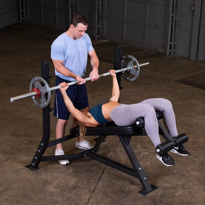 A man assisting a woman in exercising on the Body-Solid Pro Clubline Decline Olympic Bench SODB250, as she performs a bench press with a barbell in a gym setting.