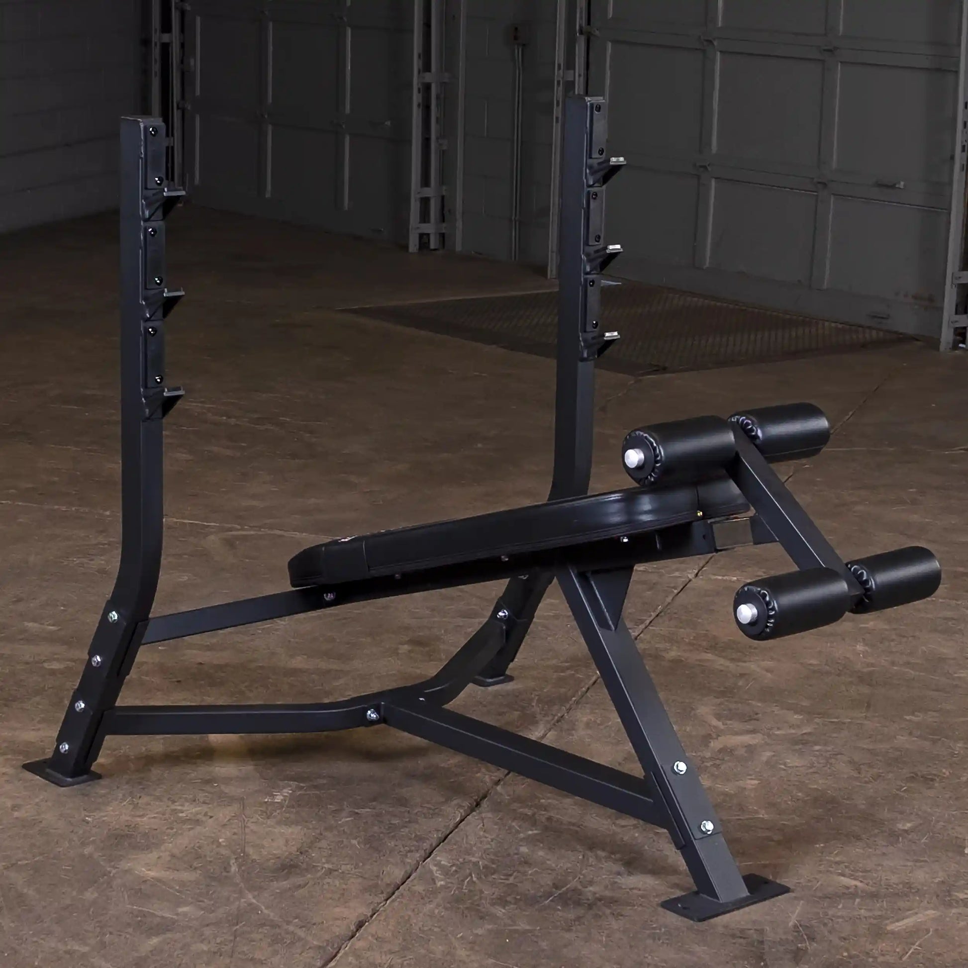Body-Solid Pro Clubline Decline Olympic Bench SODB250, showcasing a sturdy black frame with adjustable height settings and leg rollers, set in a gym environment.