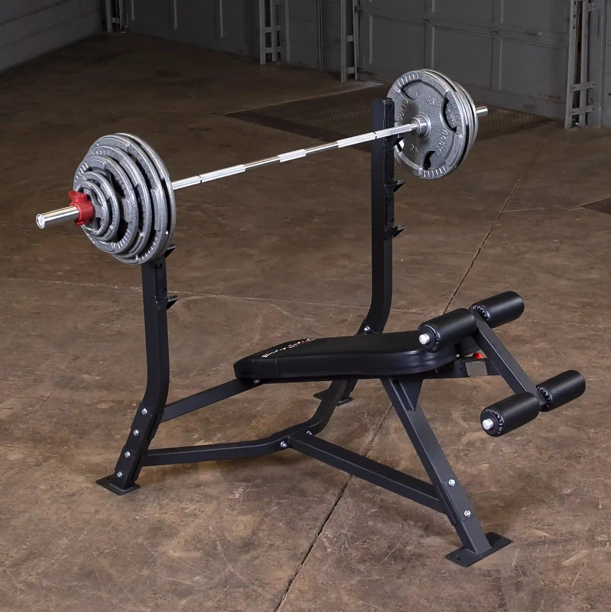 Image of a Body-Solid Pro Clubline Decline Olympic Bench SODB250 with a barbell and weight plates, set against a textured floor in a gym environment.