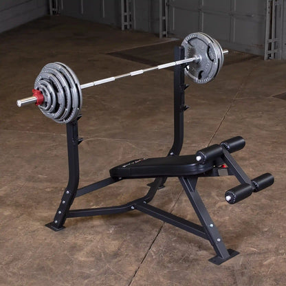 Image of a Body-Solid Pro Clubline Decline Olympic Bench SODB250 with a barbell and weight plates, set against a textured floor in a gym environment.