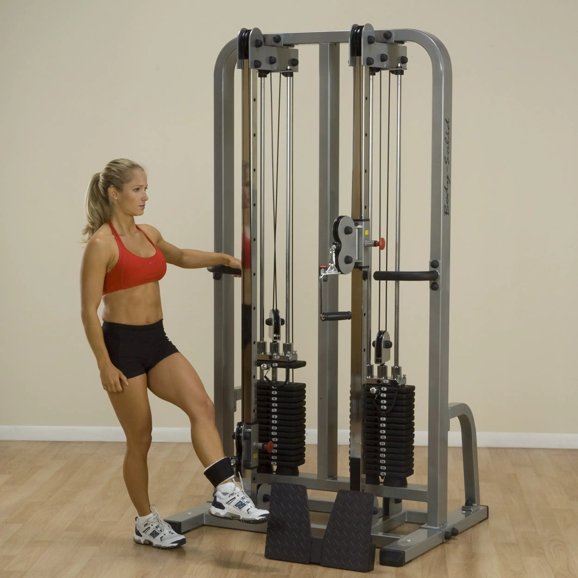 A woman in a red sports bra and black shorts using the Body-Solid Pro Clubline Dual Cable Column SDC2000G2 for exercise, with weights visible on the machine.