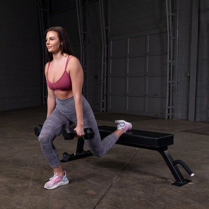 A woman performing a single-arm dumbbell row on a Body-Solid Pro Clubline Flat Bench SFB125 in a gym setting. She is wearing a pink sports bra and gray leggings.