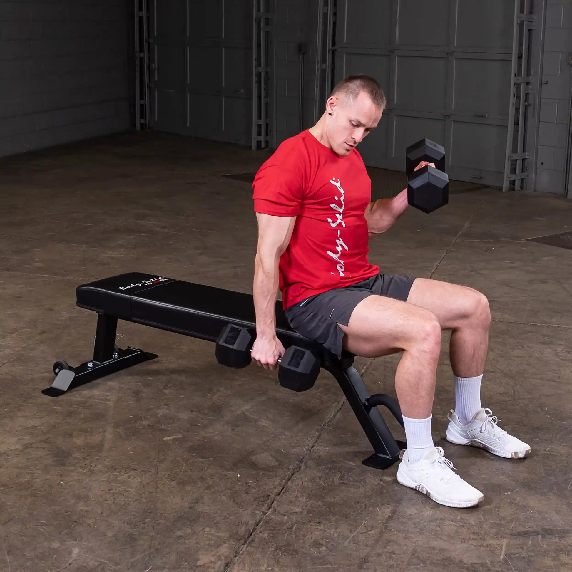 A person sitting on the Body-Solid Pro Clubline Flat Bench SFB125, performing a dumbbell exercise, wearing a red shirt and shorts in a gym setting.
