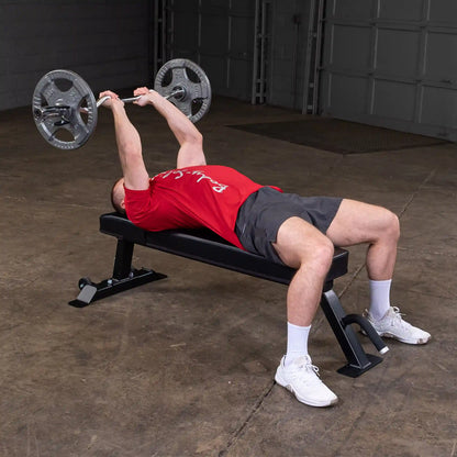 A person performing a bench press exercise on the Body-Solid Pro Clubline Flat Bench SFB125, lifting a barbell with weights in a gym setting.