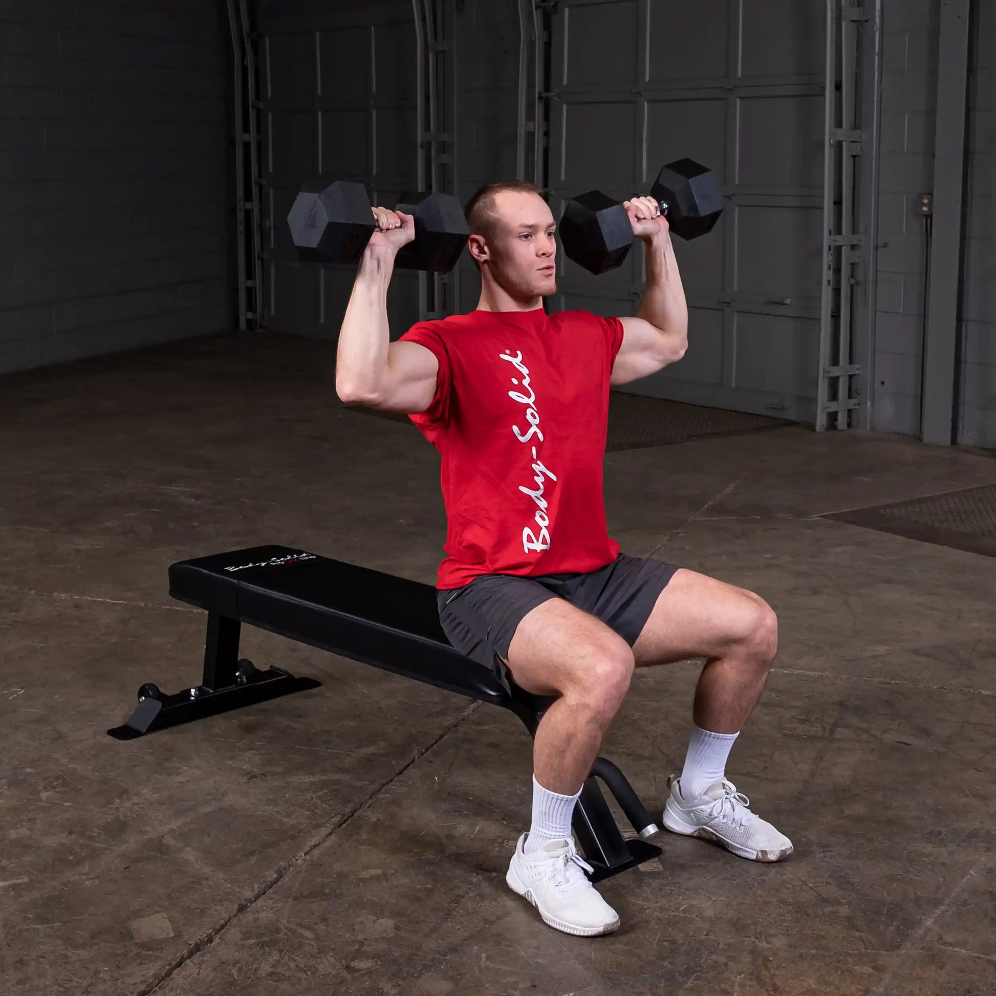 A person seated on a Body-Solid Pro Clubline Flat Bench SFB125, performing an overhead dumbbell press with weights in both hands.