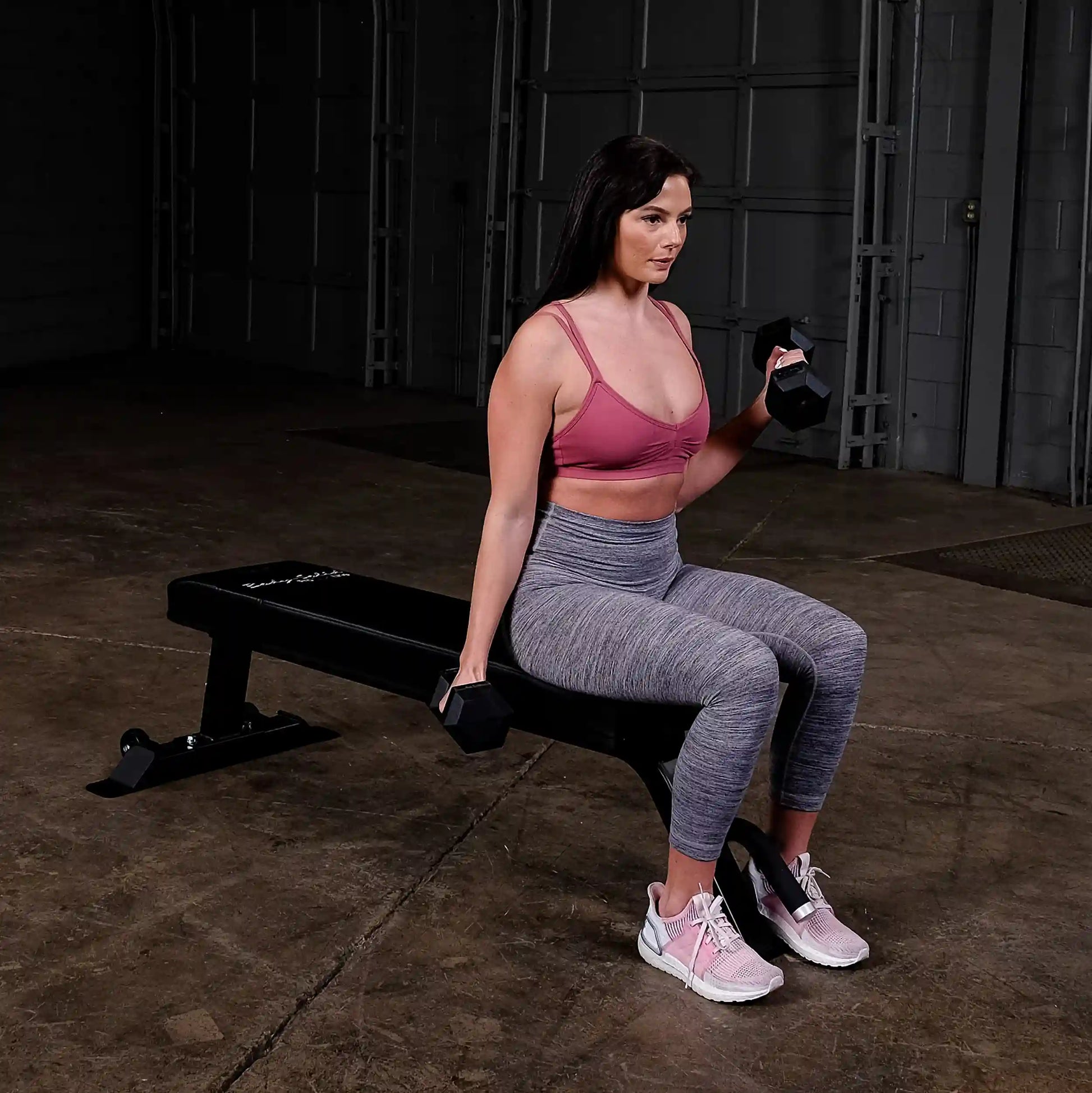 A woman sitting on a Body-Solid Pro Clubline Flat Bench SFB125, holding dumbbells in each hand, wearing a pink sports bra and gray leggings, in a gym setting.