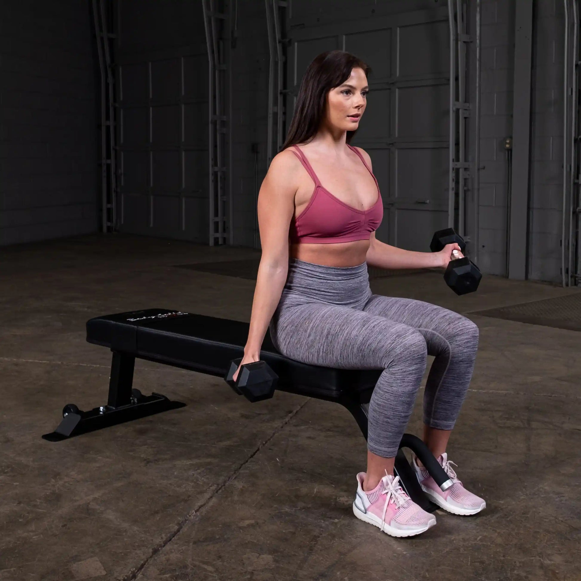 A woman sitting on the Body-Solid Pro Clubline Flat Bench SFB125, holding dumbbells while preparing for a workout in a gym setting.