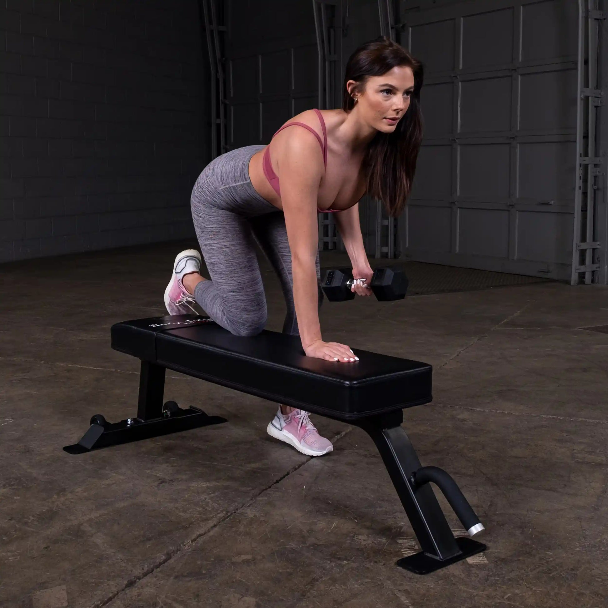 A woman exercising on the Body-Solid Pro Clubline Flat Bench SFB125, performing a dumbbell row with one knee on the bench.
