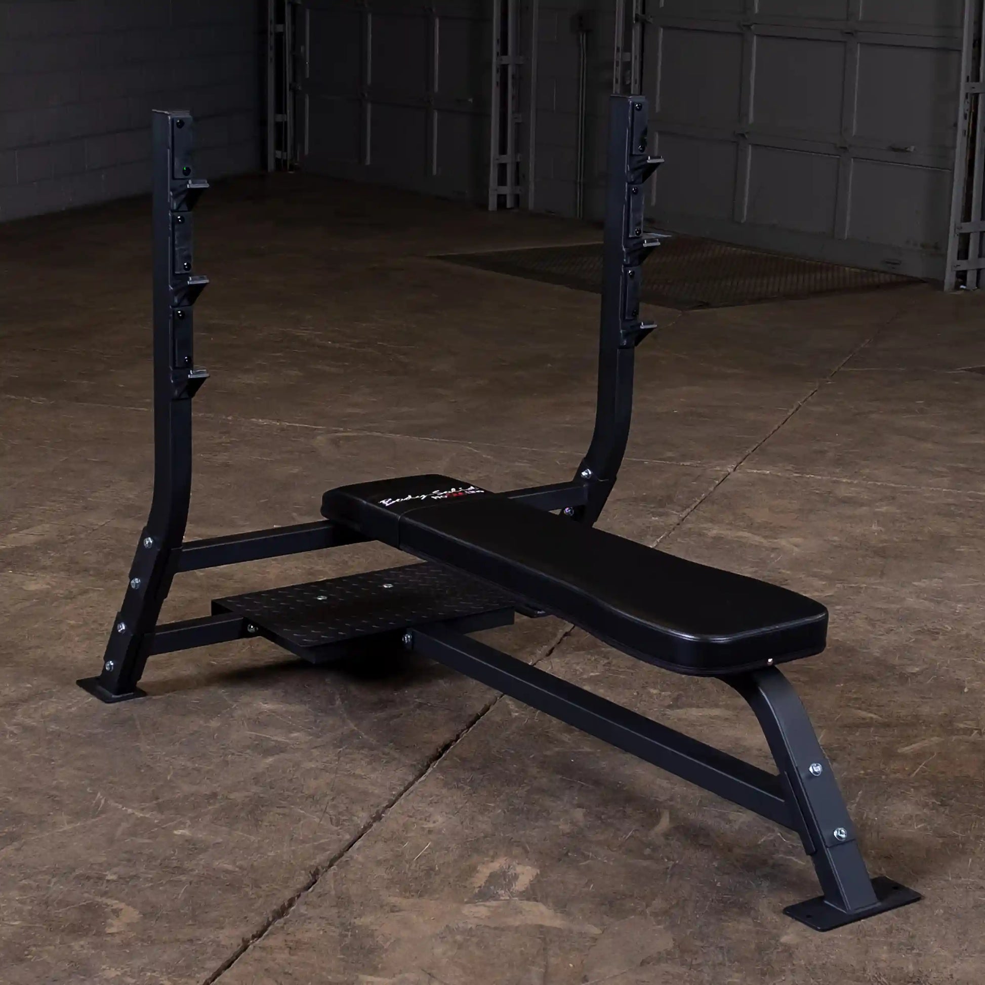 Body-Solid Pro Clubline Flat Olympic Bench SOFB250 positioned in a gym setting on a concrete floor, featuring a sturdy black frame and padded bench surface.