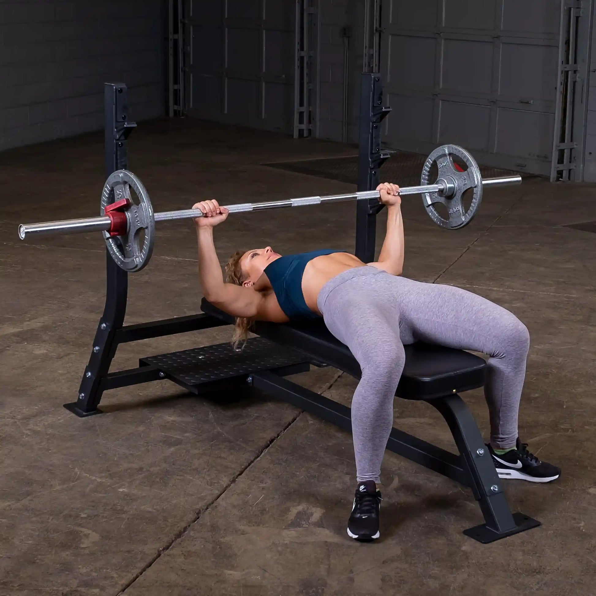 A person performing a bench press on the Body-Solid Pro Clubline Flat Olympic Bench SOFB250, featuring a barbell with weights, in a gym setting.
